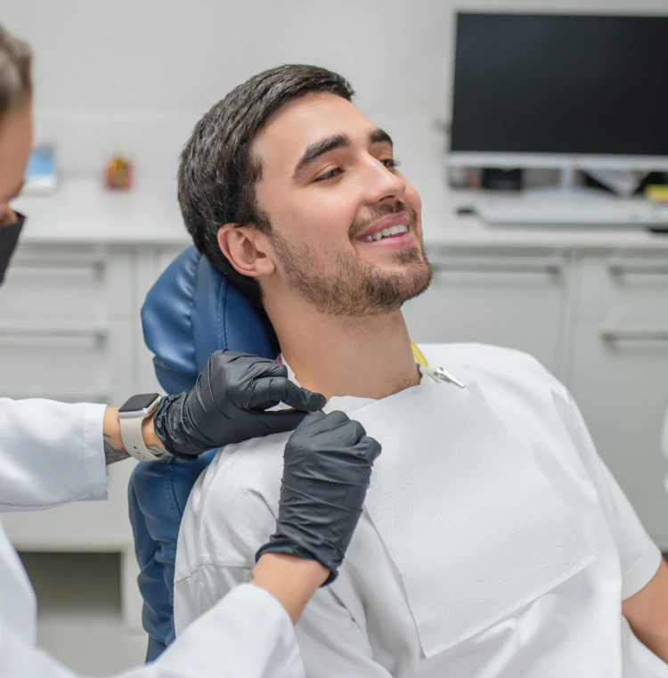 A man sits and smiles in a dental chair while a dental professional wearing black gloves attaches a protective bib around his neck in a modern dental office.