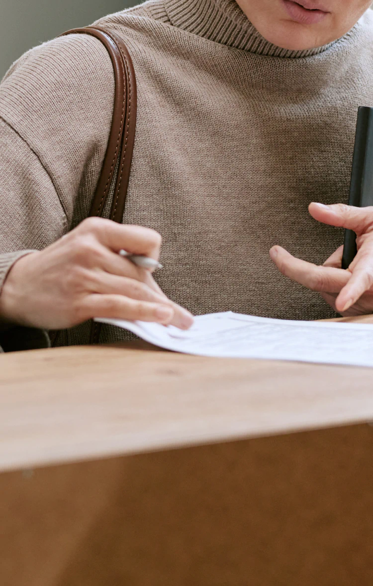 Desktop_About_Us_Insurance_Information A person wearing a beige sweater is signing documents at a wooden desk, holding a pen in one hand and a folder in the other. Their face is partially visible.