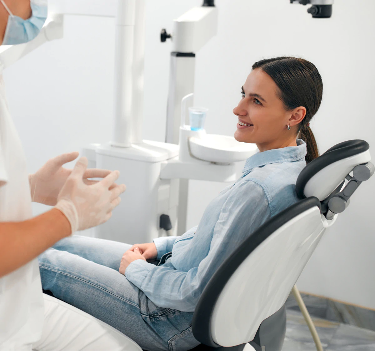 Desktop_About_Us_Precision_Guided A woman sits in a dental chair, smiling at a dentist who is wearing gloves and talking to her in a bright, modern dental office.