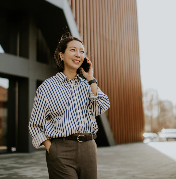 Smiling woman in a striped shirt talks on her phone while walking outside near a modern building with vertical brown panels.