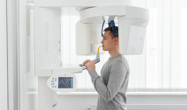 Desktop_Home_Advanced_Low_Radiation A man stands in a dental imaging machine, holding handles and biting a yellow mouthpiece for a panoramic dental X-ray scan in a bright medical room.