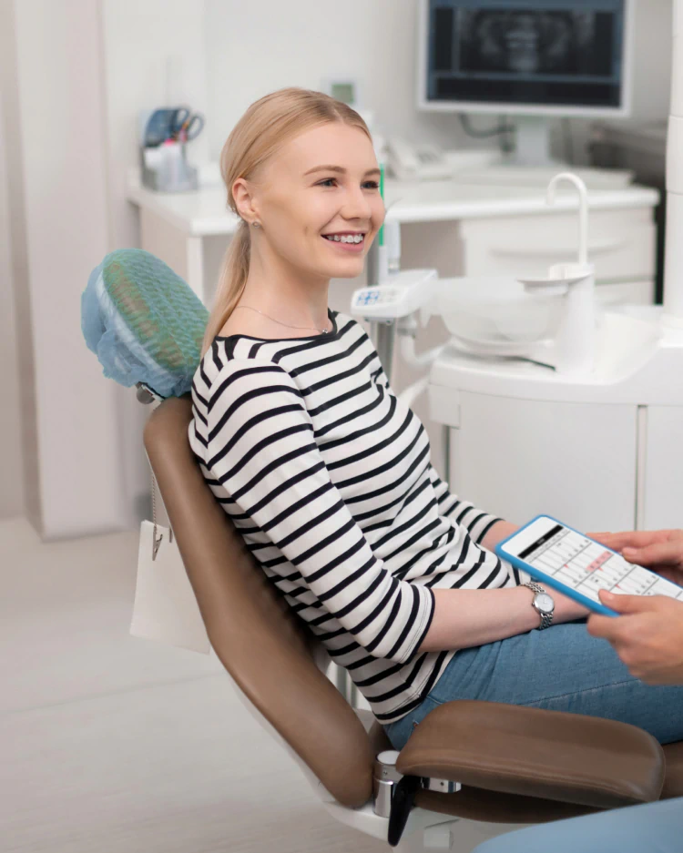 Desktop_Home_Advanced_Procedures A woman in a striped shirt smiles while sitting in a dental chair, holding a clipboard. Dental equipment and a computer screen are visible in the background.