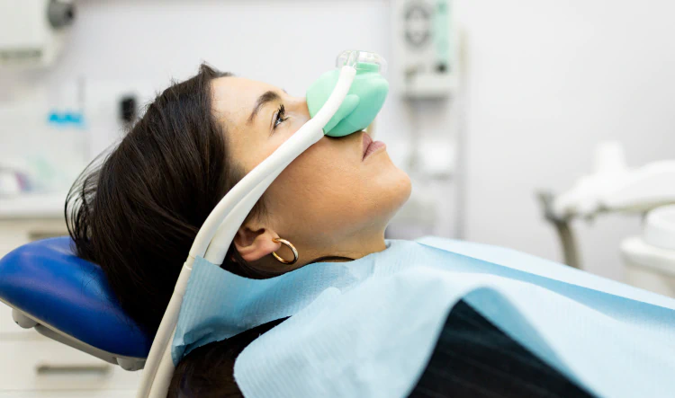 Desktop_Home_Comfortable_Sedation A woman lies in a dental chair wearing a nasal mask for sedation, with a blue bib over her chest in a dental clinic setting.