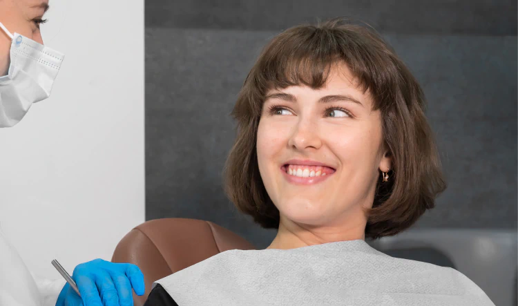 Desktop_Home_Compassionate_Highly_Attentive A woman with short brown hair smiles while sitting in a dental chair, wearing a dental bib. A dental professional in a mask and gloves is next to her, holding a dental tool.