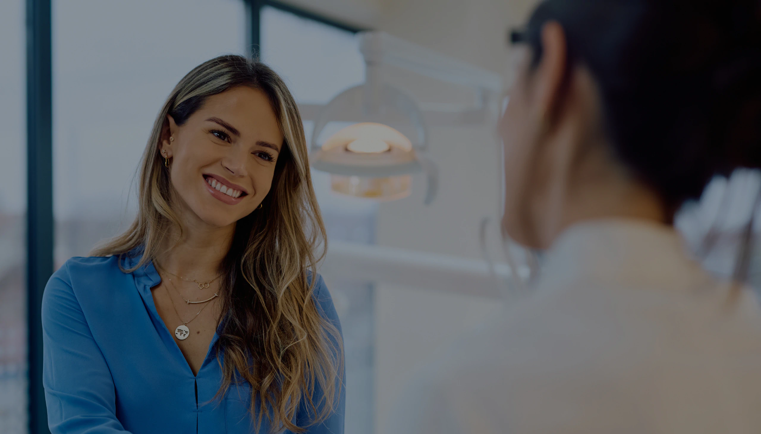 Desktop_Home_Hero A woman with long hair in a blue shirt smiles while talking to another person in a bright, modern office setting.