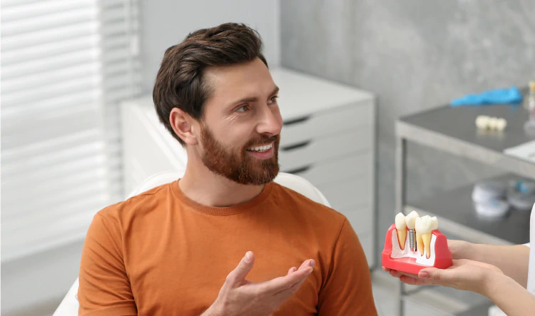 Desktop_Home_Personalized_Treatment_Plans A man with a beard smiles while sitting in a dental clinic, gesturing toward a dental model with implants and teeth that a dentist is holding.