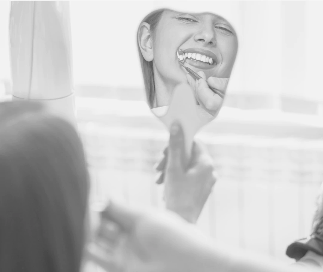 Desktop_Home_Proudly_Serving A person smiles while looking into a handheld mirror, with another hand using dental tools near their mouth, suggesting a dental checkup or cleaning.