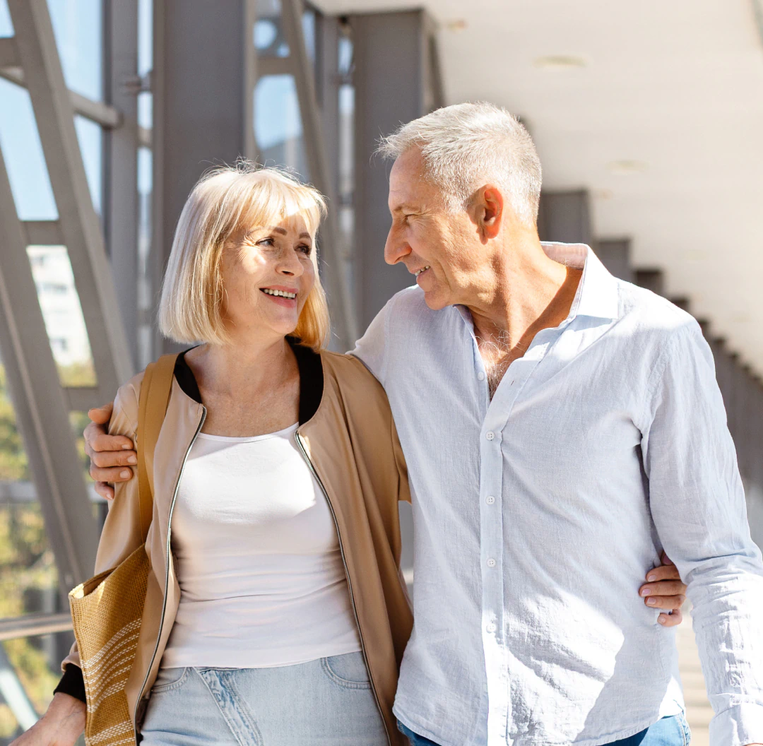 Desktop_Home_Raising_the_Standard An older couple smiles at each other while walking arm in arm on a sunny day, enjoying each others company in an outdoor corridor with lots of natural light.