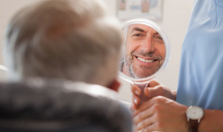 Desktop_Home_Recovery_Focused An older person looks into a small handheld mirror, which reflects the smiling face of a middle-aged man. Another person, partially visible, holds the mirror for them.