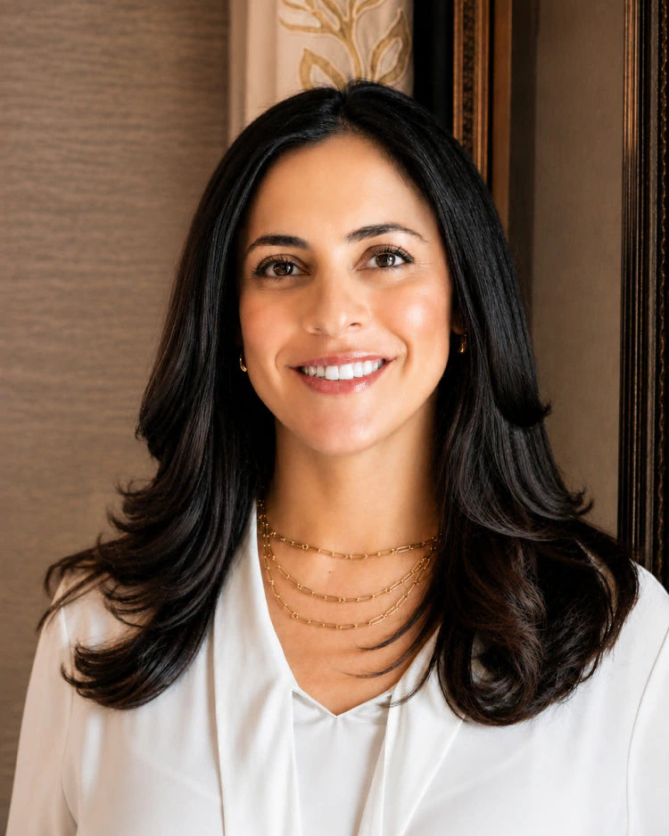 Desktop_Home_Surgical_Expertise A woman with long dark hair, wearing a white blouse and layered gold necklaces, smiles at the camera. She stands in front of a neutral background with a decorative curtain and a framed edge visible.