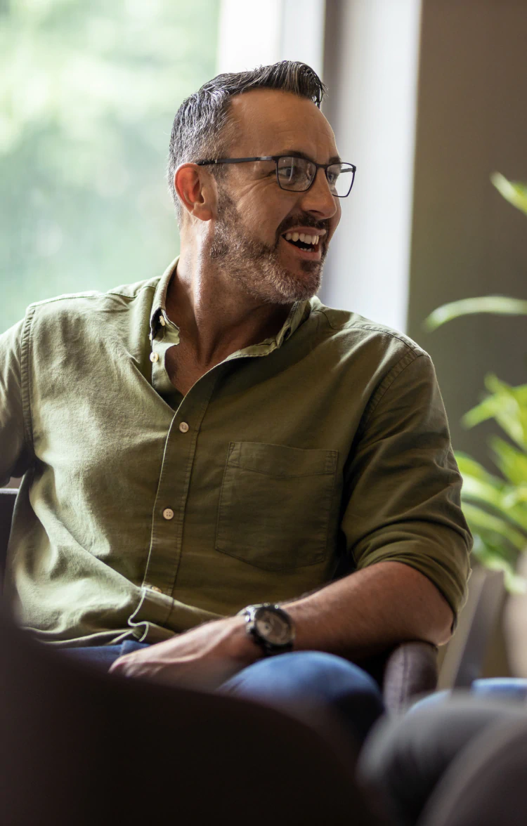 Desktop_Home_The_Dailey_Center_Experience Smiling man with short hair, a beard, and glasses sits on a chair, wearing a green button-up shirt and watch, with a blurred green plant and window in the background.