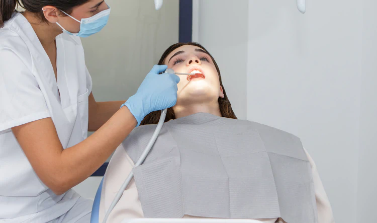 Desktop_Home_Trauma_Trained A dentist, wearing gloves and a mask, examines a patient’s open mouth with dental tools in a brightly lit clinic. The patient sits reclined, covered with a gray protective bib.