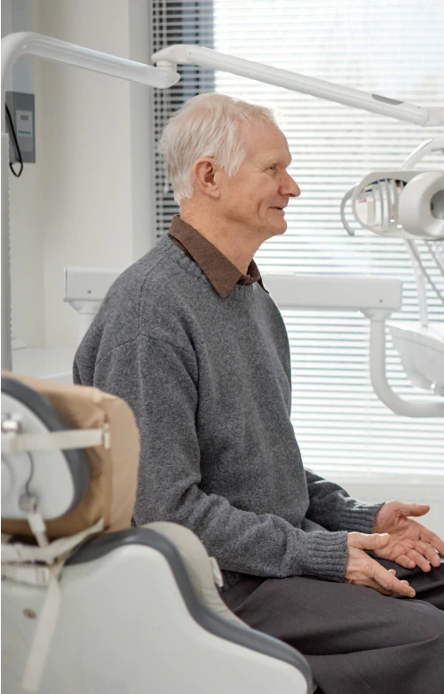 Desktop_Meet_the_Doctor_Drop_Downs An older man with gray hair sits in a dentists office, smiling and wearing a gray sweater. Dental equipment and a chair are visible in the background.