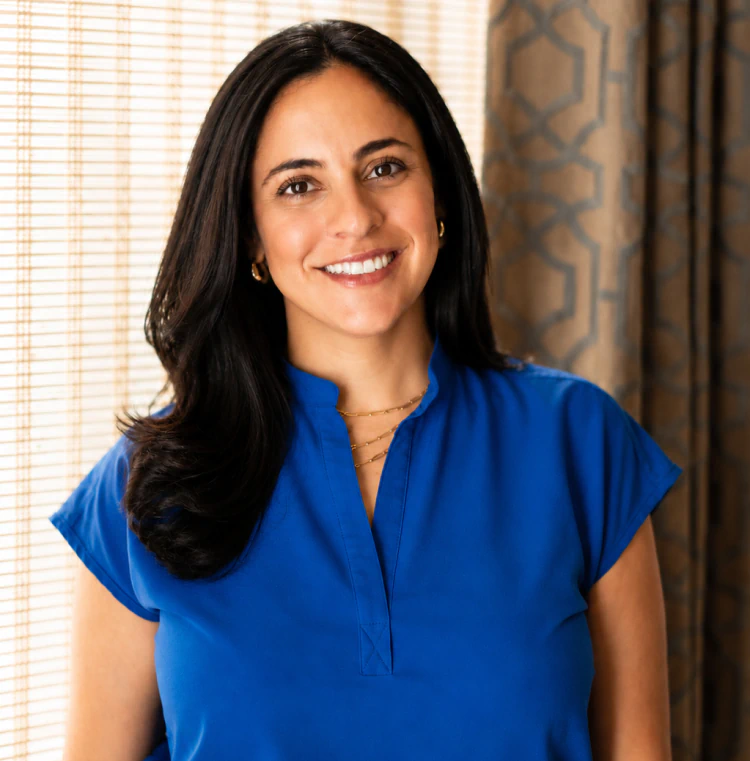 Woman with long black hair wearing a bright blue blouse smiles, standing indoors in front of a window with bamboo blinds and patterned curtains.