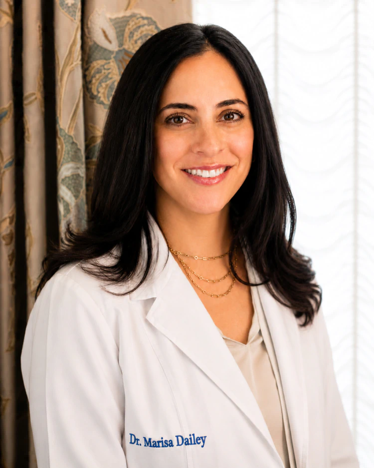 Desktop_Meet_the_Doctor_Marisa_Dailey_DDS A woman with long dark hair wearing a white lab coat and gold necklaces smiles while standing indoors in front of a curtain. The lab coat reads Dr. Marisa Dailey.