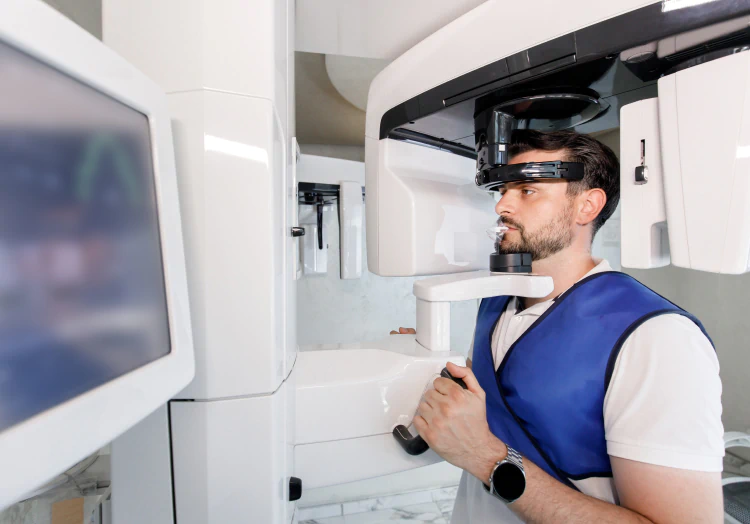A man wearing a blue lead apron stands still as a dental panoramic X-ray machine scans his head in a modern medical clinic.