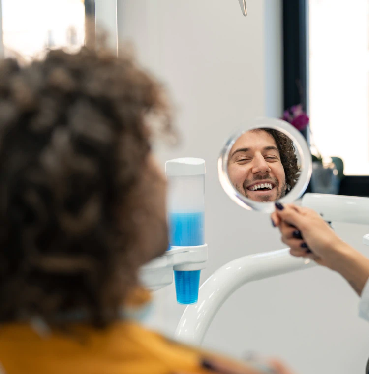 Desktop_Services_Hero A person with curly hair smiles while looking at their reflection in a round handheld mirror at a dental clinic.