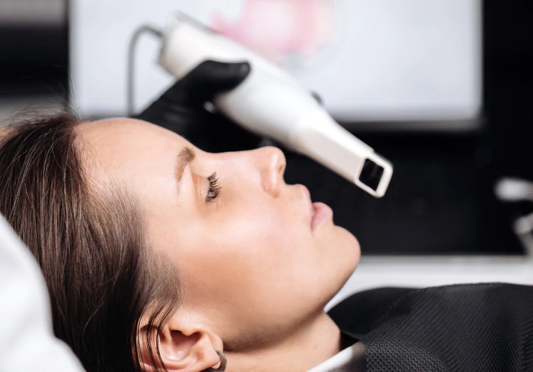 A woman lies with her eyes closed while a gloved hand holds a white handheld device to her face, possibly for a skin or facial treatment.