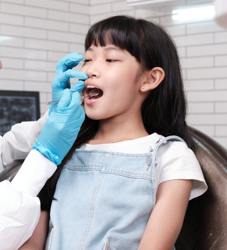 A young girl sits in a dental chair with her mouth open while a dentist wearing blue gloves examines her teeth in a brightly lit clinic.