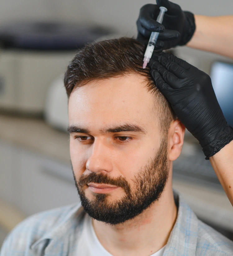 A man with a beard receives an injection in his scalp from a person wearing black gloves, possibly for a hair treatment, in a medical or clinic setting.