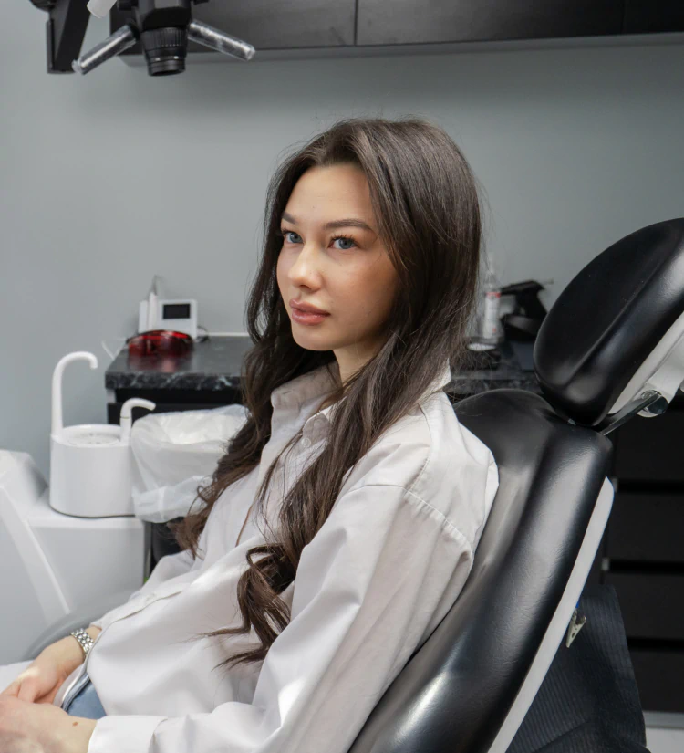 A woman with long dark hair sits in a dentist chair, looking at the camera. Dental equipment and a sink are visible in the background.