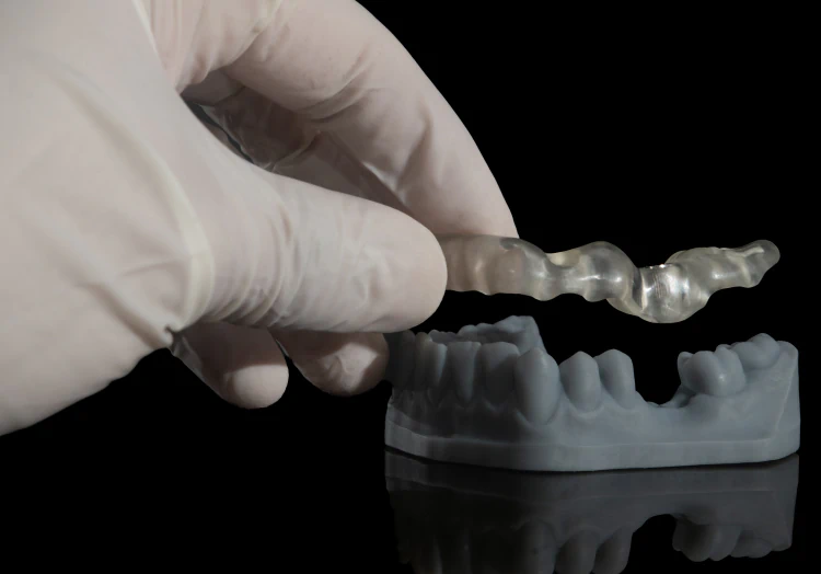 A gloved hand holds a clear dental aligner above a gray dental mold against a black background.