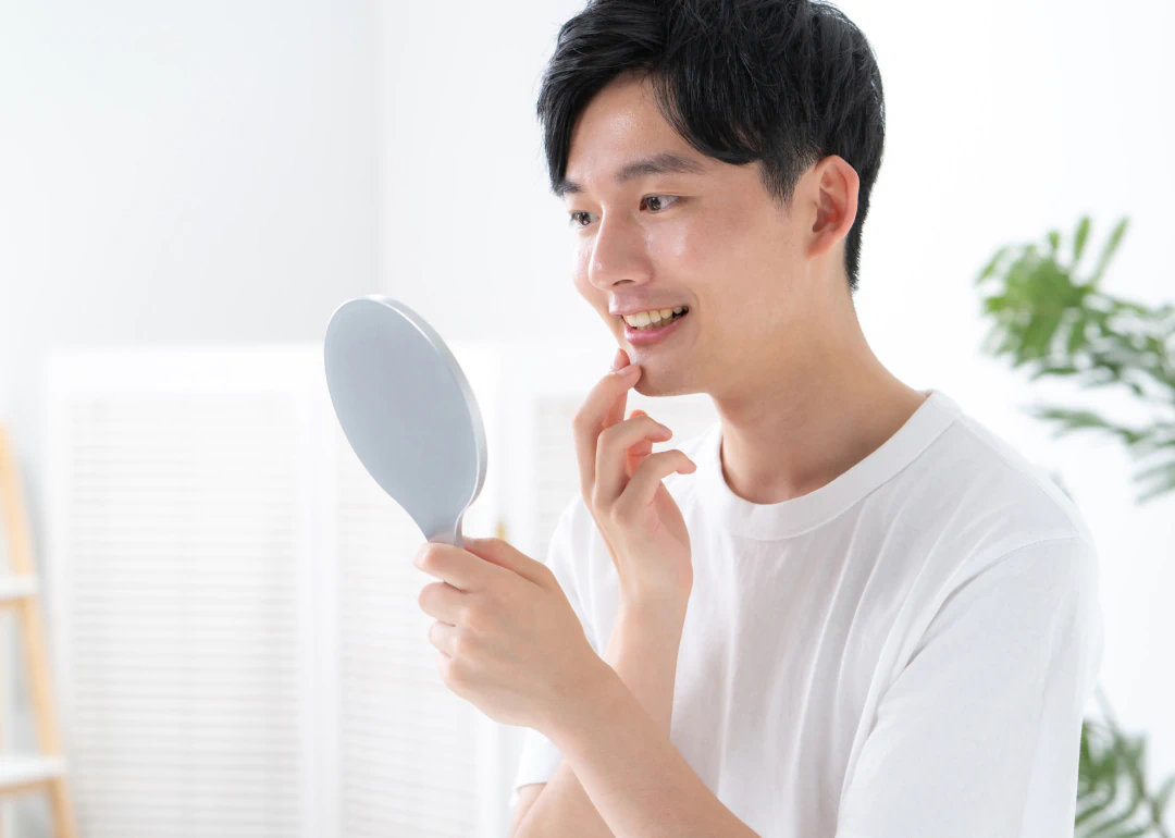A young man in a white shirt smiles while looking at his reflection in a handheld mirror, touching his face with one finger. There is a green plant in the background.