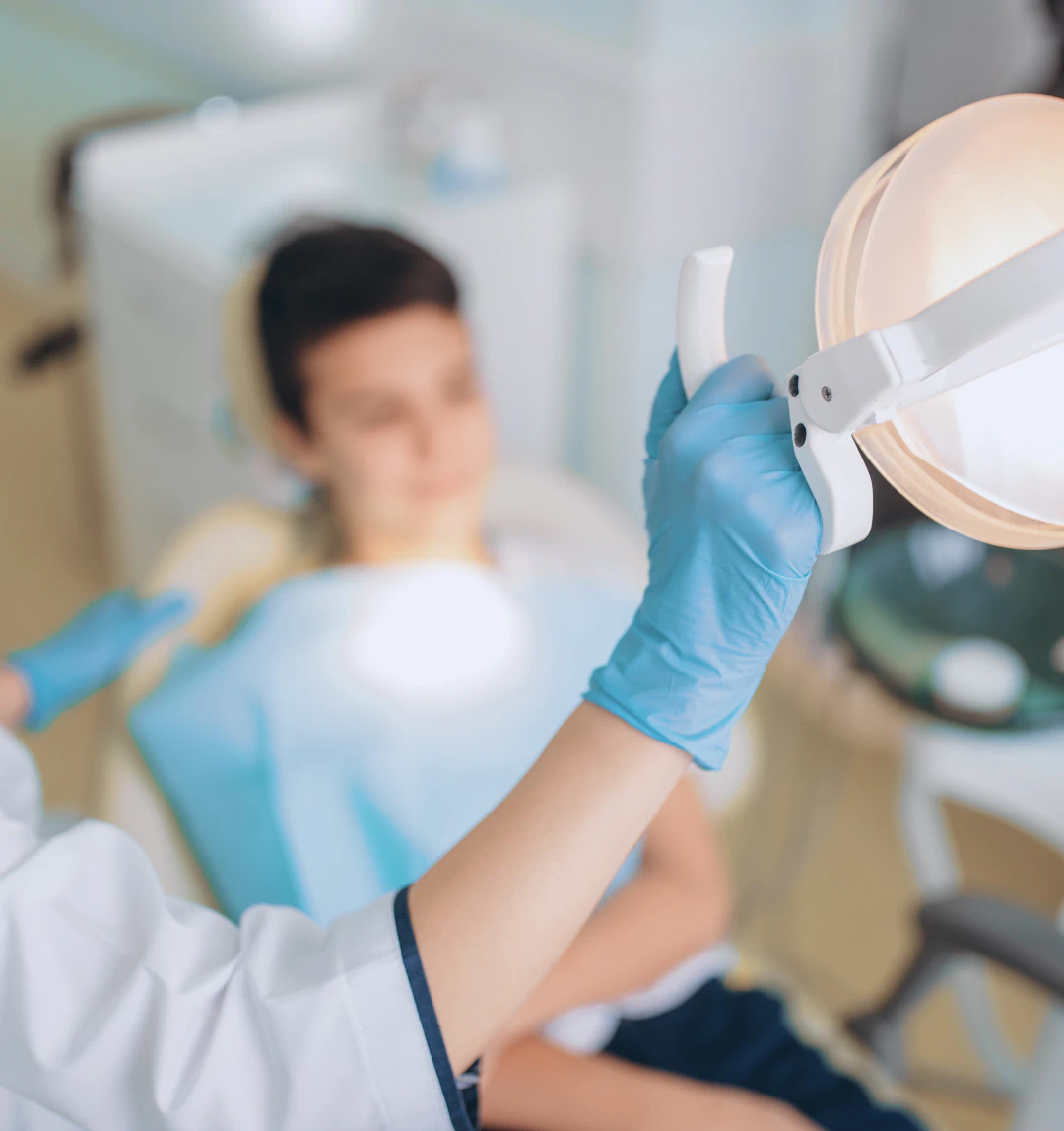 A person wearing blue gloves adjusts a dental lamp above a patient lying in a dentist’s chair. The patient is out of focus in the background, while the focus is on the gloved hand and the lamp.