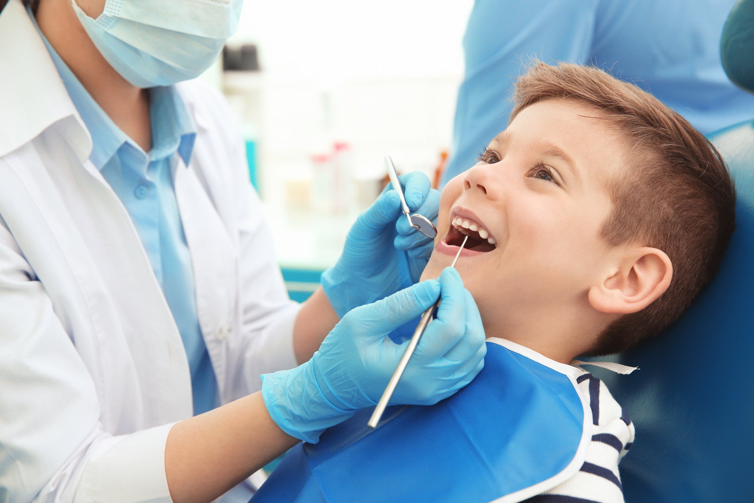 AdobeStock_161295211 v2 A pediatric dentist in a mask and blue gloves examines a young boys teeth in a dental chair in Wall Township